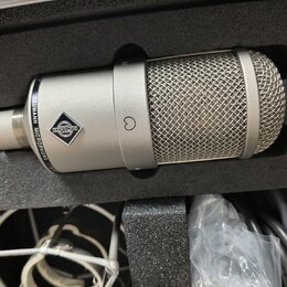 Stříbrný kondenzátorový mikrofon v černé pěnové výplni kufru. Na těle je vidět logo značky a nápis NEUMANN MICROPHONE. V dolní části kufru jsou vidět…