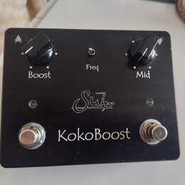 Černý kytarový efekt s nápisem KokoBoost a logem Suhr. Na horní straně jsou dva černé otočné knoflíky s popisky Boost a Mid, mezi nimiž se nachází ma…