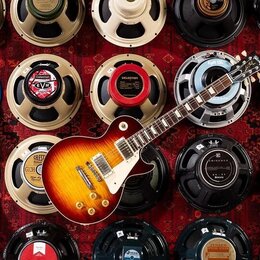 Elektrická kytara s tělem v žlutě-oranžovém gradientu (sunburst) a bílým pickguardem leží napříč řadami kytarových reproduktorů. Na hlavě kytary je v…