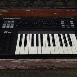 Černý MIDI kontrolér s klaviaturní klávesou. Na horním panelu je nápis KOMPLETE KONTROL S25 a v pravém horním rohu označení MK1. Zařízení obsahuje ně…