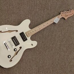 Bílá polodublová elektrická kytara s přírodním hřidlem a hlavou. Na hlavě je nápis "Squier STARSCASTER". Tělo má dva humbuckery, dva ovládací knoflík…