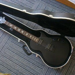 Černá elektrická kytara s tvarem singlecut v otevřeném černém tvrdém kufru. Na hlavě je viditelné logo ESP. Kytara má dva humbuckery, dvě kontrolní k…