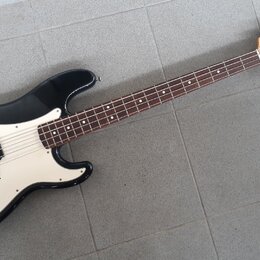 Černá elektrická basová kytara s bílým pickguardem a hnědým hmatníkem. Na hlavě nástroje je viditelné logo Squier a nápis Precision Bass. Povrch kyta…