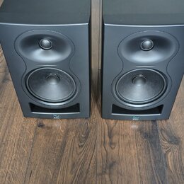 Dva černé regálové reproduktory umístěné na dřevěné podlaze. Každý reproduktor má v horní části výšký jednotku a ve spodní části basový reproduktor.…
