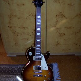 Elektrická kytara s tónem sunburst, bílým pickguardem a dvěma humbuckery. Na hlavě je nápis BaCha a pod ním logo ve tvaru stylizovaného písmena V. Hр…