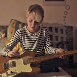 Dítě v proužkovaném tričku drží v rukou elektrickou kytaru s přírodním dřevěným povrchem těla. Kytara má bílý pickguard, dva snímače a tři ovládací k…