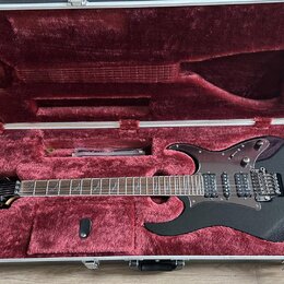 Černá elektrická kytara v hliníkovém kufru s červeným plyšovým vnitřkem. Na hlavě nástroje je bílý nápis Ibanez. Kytara má dva humbuckery, tremolo mo…
