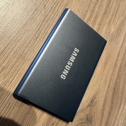 Na fotografii je modrý obdélníkový předmět s kovovým povrchem, který leží na dřevěném stole. Uprostřed je bílý nápis SAMSUNG. Povrch předmětu vypadá…