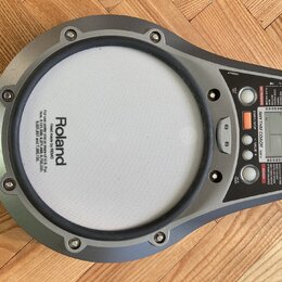 Šedý elektronický tréninkový pad značky Roland s bílou úderovou plochou. Na ovládacím panelu je digitální displej, nápis RHYTHM COACH a modelové ozna…