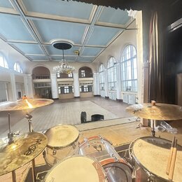 Fotografie zachycuje bicí soupravu umístěnou na pódiu v prostorné sále. Na činelech je vidět nápis Paiste a na blbáku logo REMO. Souprava má průhledn…