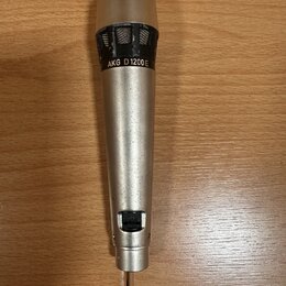 Stříbrný dynamický mikrofon s kovovým tělem a mřížkou. Na horní části je černý nápis AKG D1200E. Povrch těla vykazuje drobné škrábance a známky opotř…