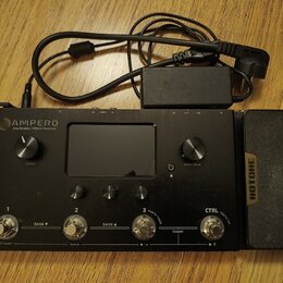 Černý kytarový procesor s nápisem AMPERO Amp Modeler / Effects Processor. Zařízení má displej, dva otočné knoflíky označené VOLUME a MENU / VALUE, tř…