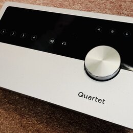Stříbrný audio rozhraní s černým ovládacím panelem a velkým kovovým otočným knoflíkem. Na šasi je vlevo logo a nápis APOGEE a vpravo nápis Quartet. N…