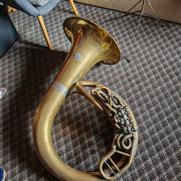 Zlatý mosazný barytonsaxofon položený na koberci. Na zvonu je viditelné kulaté logo, které je však nečitelné. Povrch nástroje vykazuje známky opotřeb…