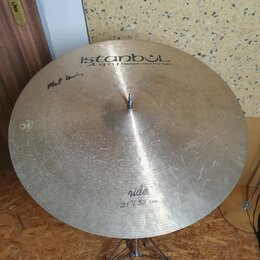 Kruhové činely typu ride z mosazného kovu s viditelnými stopami používání a drobnými škrábancemi. Na povrchu je nápis istanbul Agop Handmade cymbals…