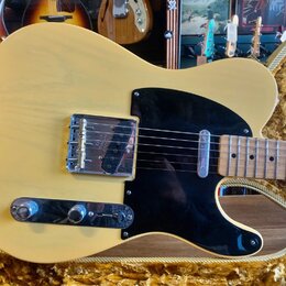 Kytara typu Telecaster v krémové barvě s černým pickguardem a hnědým javorovým hmatníkem. Na kovovém krytu snímače je nečitelný nápis. Nástroj leží v…