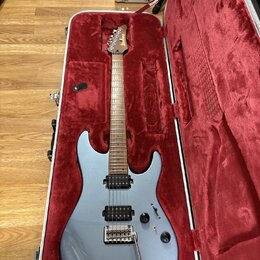 Stříbrná elektrická kytara v otevřeném tvrdém kufru s červeným vnitřkem. Na hlavě hředu je bílý nápis Ibanez. Kytara má dva humbuckery, dva ovládací…