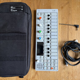 Na dřevěném stole je bílý syntezátor OP-1 s řadou ovládacích knoflíků v různých barvách, displejem a tlačítky. Vedle něj je černý textilní přepravní…