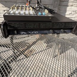 Na černém kovovém mřížkovém povrchu stojí černý rackový zesilovač s nápisem Professional Power Amplifier QSA 1000 a logem Q SOUND. Na zesilovači je u…