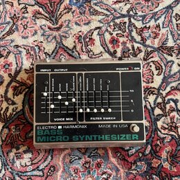 Stříbrný efektový pedál s černým ovládacím panelem. Na spodní části je nápis ELECTRO HARMONIX MADE IN USA BASS MICRO SYNTHESIZER. Panel obsahuje něko…