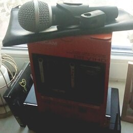 Na černé podložce leží dynamický mikrofon s kovovou mřížkou a černým tělem. Pod ním je červená krabice s nápisy CAM a DR-V1HD. Vedle ní stojí černá k…