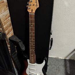 Elektrická kytara s tónem sunburst a bílým pickguardem. Na hlavě hředu je viditelné logo Fender a nápis STRATOCASTER. Kytara má hnědý hmatník a je op…