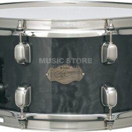 Černý snare drum s kovovým obručováním a chromovaným mechanismem. Na koruse je zlatý oválný štítek s nápisy „TAMA“, „Custom Superior“ a podpisem, kte…