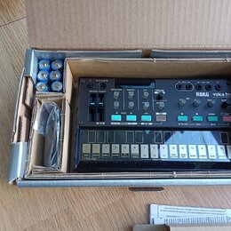 V kartonové krabici se nachází černý syntezátor s nápisem KORG volca fm. Přístroj má bílé ovládací prvky, posuvníky a řadu 16 tlačítek v dolní části.…