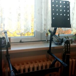 Černý skladací notový stojan s perforovanou deskou. Stojan je umístěn v interiéru u okna, vedle něj je vidět hlava kytary. Na fotce nejsou žádné čite…