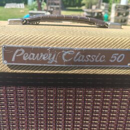 Detail kytarového komba potaženého béžovou texturou s hnědým koženým úchem. Na přední straně je kovový štítek s nápisem Peavey Classic 50. Povrch ští…