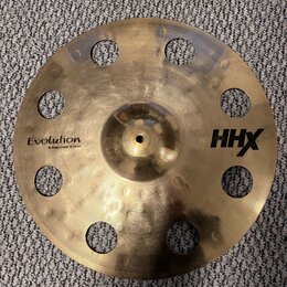 Zlatý kovový činel s otvory v okruhu kolem středu. Na povrchu jsou nápisy Evolution, O-Zone Crash 15" 40 cm a HHX. Povrch vykazuje jemné rýhy a drobn…