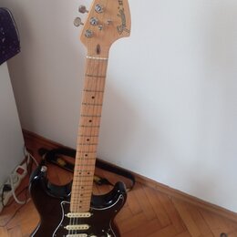 Černá elektrická kytara typu Stratocaster s jasným dřevěným hředníkem a krčkem. Na hlavě je vidět nápis Fender STRATOCASTER. Kytara má tři snímače, b…
