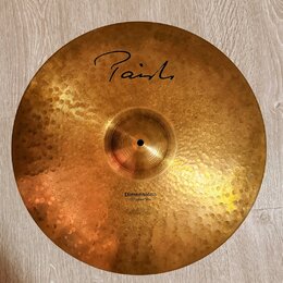 Zlatavý kovový činel s kovaným povrchem. Na horní části je černý nápis Paiste a ve spodní části text Dimensions Hand Hammered Ride. Povrch vykazuje d…
