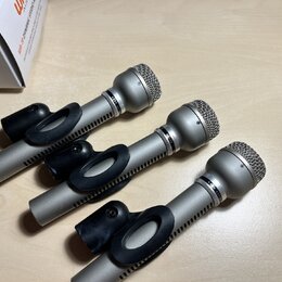 Tři stříbrné dynamické mikrofony s kovovými mřížkami a černými plastovými držáky. Na tělech mikrofonů jsou viditelné rýhované prstence, ale žádná zna…