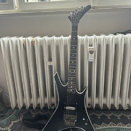 Černá elektrická kytara s ostře hranatým tvarem těla a špičatou hlavou. Na hmatníku jsou vidět bílé polohové značky a na těle jsou dva humbuckery, ov…