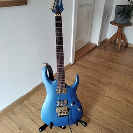 Modrá elektrická kytara s kovovými zlatými komponenty, umístěná na černém stojanu. Na hlavě je viditelné logo Ibanez. Kytara má dva humbuckery, tremo…