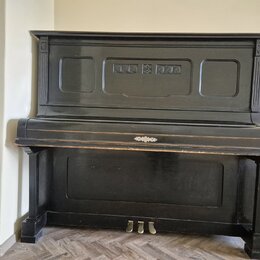 Černý piano v přímém záběru zepředu. Nástroj má zdobný ornament uprostřed klaviaturního víka a tři mosazné pedály. Povrch vykazuje známky opotřebení…