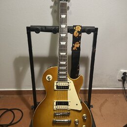 Elektrická kytara s žlutým gradientním lakem v provedení singlecut, umístěná na černém stojanu. Na hlavě je nápis Epiphone a logo Les Paul Custom. Ky…