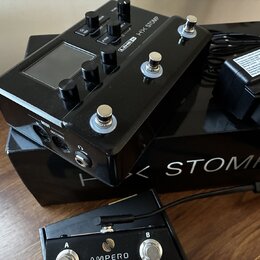 Černý efektový pedál s nápisem LINE 6 HX STOMP, vybavený displejem, několika ovládacími knoflíky a třemi stoupakovými spínači. Nástroj leží na černé…
