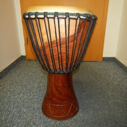 Dřevěný buben typu djembe s hnědým povrchem a světlou koženou membránou. Tělo nástroje je v horní části opletené černým šňůrem a v dolní části má vyř…