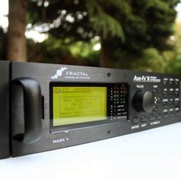 Černý rackový procesor s nápisy FRACTAL AUDIO SYSTEMS, Axe-Fx II a MARK II. Zařízení má vprostřed displej s žlutým podsvícením a textem, ovládací kno…