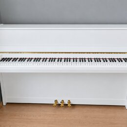 Bílé digitální piano s klaviatura a uzavíracím víkem. Na levé straně nad klávesami je viditelné kulaté logo, které je však nečitelné. Nástroj má zlat…