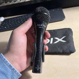 Černý dynamický mikrofon s mřížkou, v držáku. Na těle je bílý nápis AUDIX a pod ním OM-3xb, v dolní části je nápis MADE IN USA a symbol CE. V pozadí…