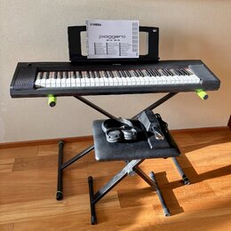 Černý digitální klavír značky YAMAHA umístěný na černém X-stojanu. Na klavíru je nápis YAMAHA a v držáku za ním je manuál s textem piaggero NP-35 NP-…