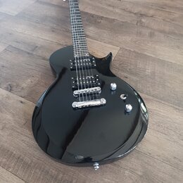 Černá elektrická kytara s tvarem připomínajícím model Les Paul. Nástroj má dva humbuckery, Tune-o-matic můstek a dvě ovládací knoflíky. Povrch je les…