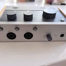Audio rozhraní v šedém provedení s dřevěnými bočními panely. Na horní straně jsou tři černé otočné ovladače s nápisy GAIN 1, GAIN 2 a MONITOR, v horn…