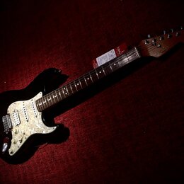 Černá elektrická kytara typu Stratocaster s bílým perleťovým pickguardem. Na hlavě hředu je viditelné logo Fender. Kytara má tmavý hmatník a na krku…