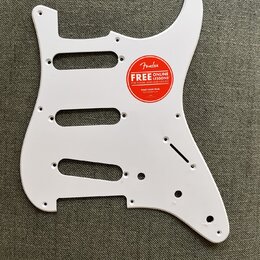 Bílá pickguard pro elektrickou kytaru s výřezy pro tři snímače. Na povrchu je nalepená červená kulatá nálepka s nápisem Fender FREE ONLINE LESSONS FO…