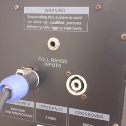 Detail zadní strany černého reproduktoru s několika konektory. Je zde vidět nápis WARNING Suspending this system should Le done by qualified persons…