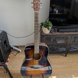 Akustická kytara s tělem v provedení sunburst (černá, hnědá a žlutá). Na hlavě je bílý nápis YAMAHA. Nástroj stojí na černém stojanu v interiéru.